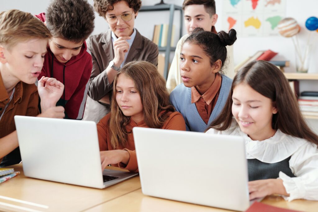 students gathered together over laptops with a teacher observing in the background