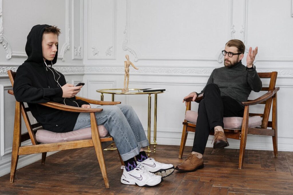 Teen male with black eye, ear pieces in ears and looking at his phone while male adult tries to make eye contact with raised hand gesture talk to a professional about suicide